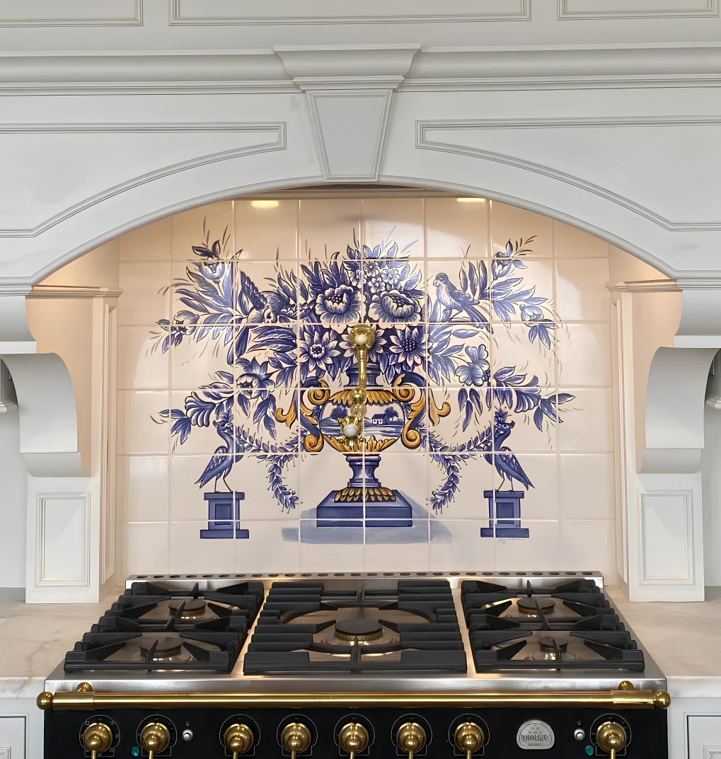 A large gas range stove with black burners and gold-tone knobs set beneath a decorative tiled backsplash featuring blue and gold floral motifs on a white kitchen wall.
